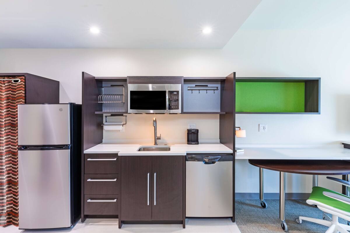 Cocina compacta con refrigerador, microondas y lavavajillas, un lavamanos pequeño y una pantalla verde en la pared.