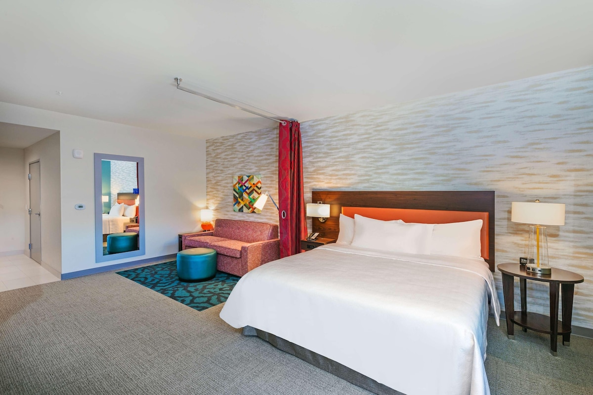 A hotel room with a bed, a desk, a chair, a small table with a lamp, and a colorful artwork on the wall.