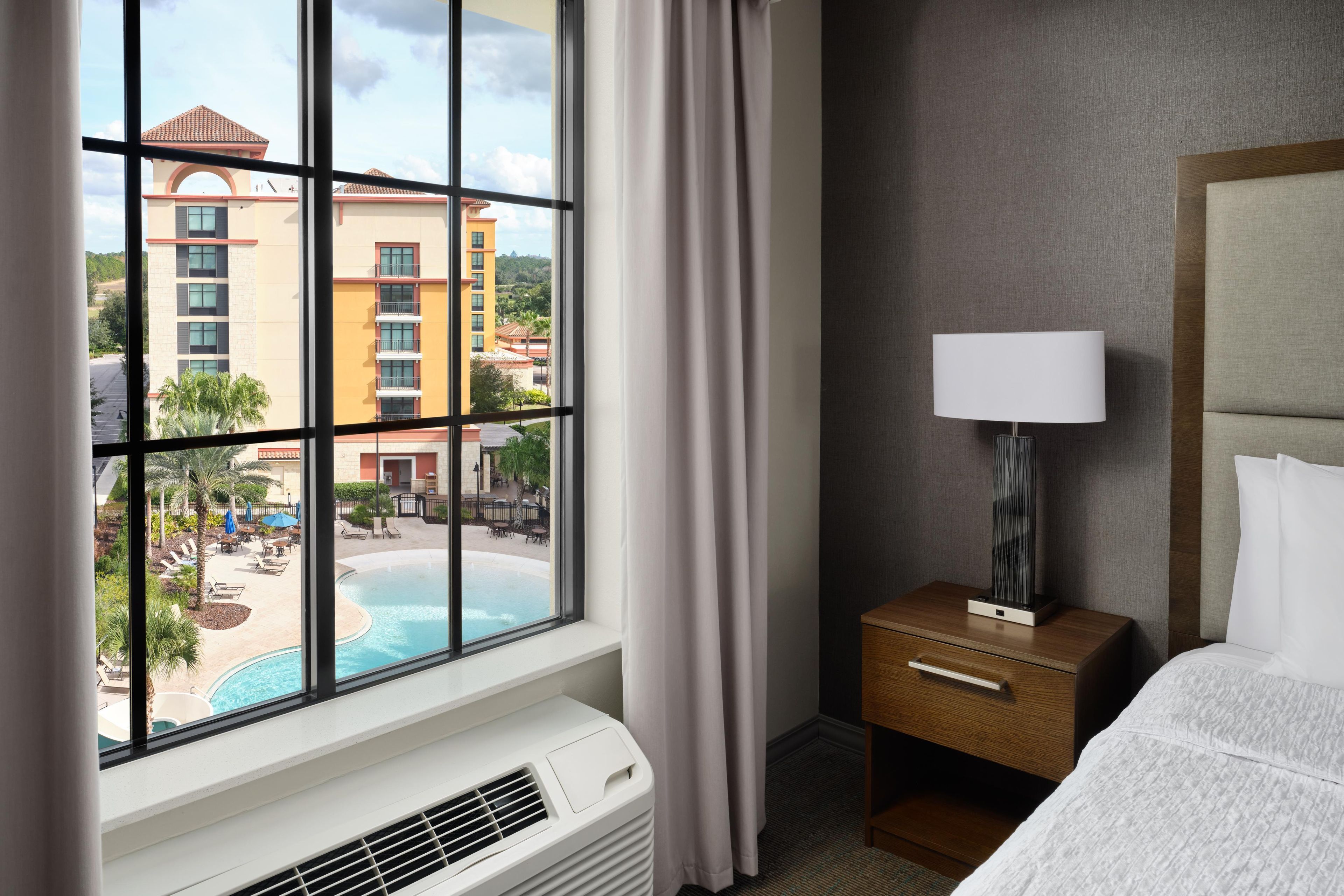 A hotel room with a large window, a bed, a bedside table with a lamp, and a wall-mounted air conditioner.