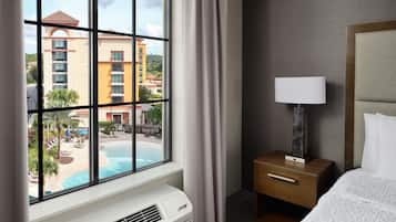 A hotel room with a large window, a bed, a bedside table with a lamp, and a wall-mounted air conditioner.