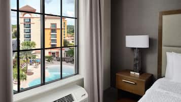A hotel room with a large window, a bed, a bedside table with a lamp, and a wall-mounted air conditioner.