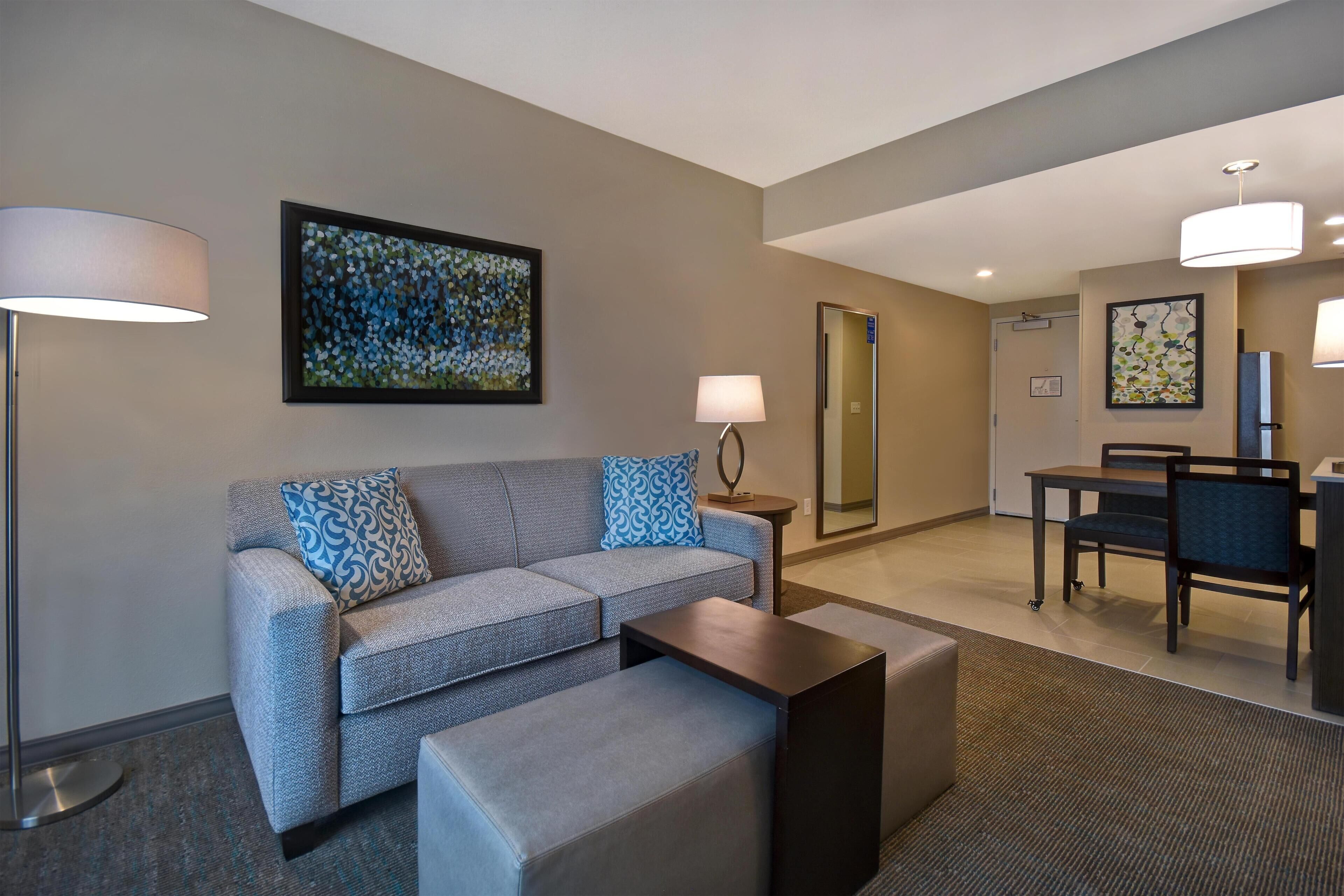 A modern hotel room with a grey sofa, a small table, a lamp, a painting, and a dining area with chairs and a table.