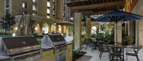 Un patio exterior con una parrilla, zona de comedor y sombrilla.