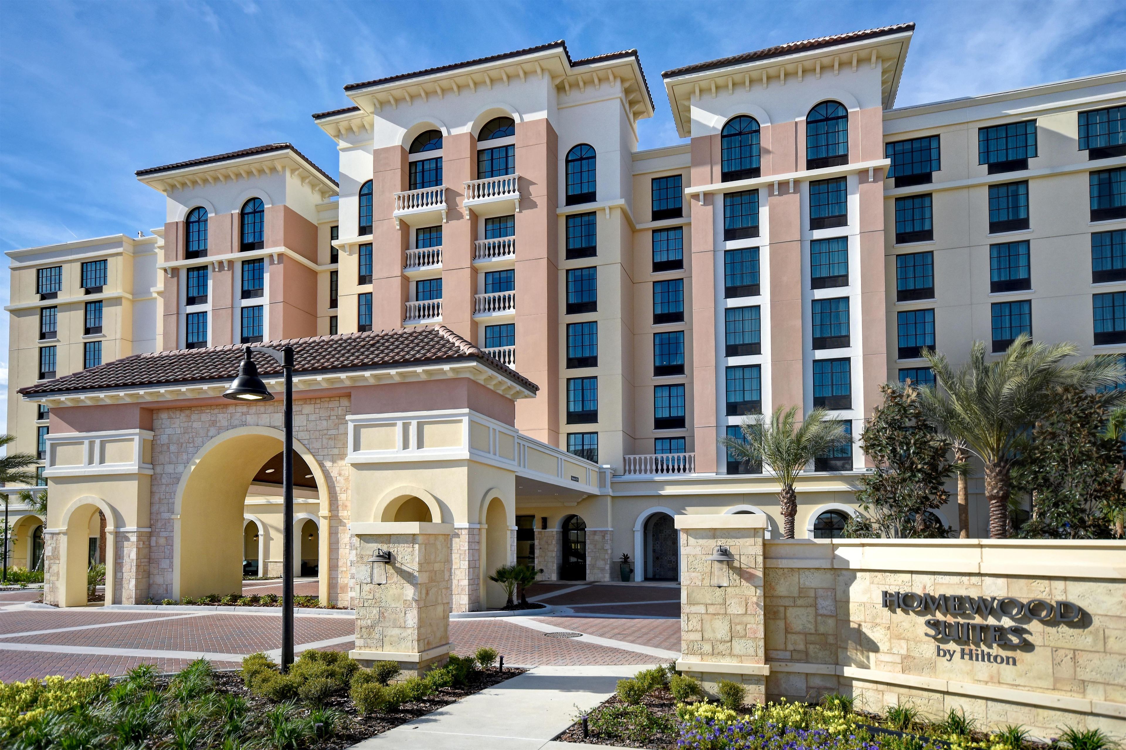 Ein mehrstöckiges Hotel mit Innenhof, das einen großen Eingangsbereich mit Bogen und eine Schild mit der Aufschrift 'Homewood Suites by Hilton' zeigt.