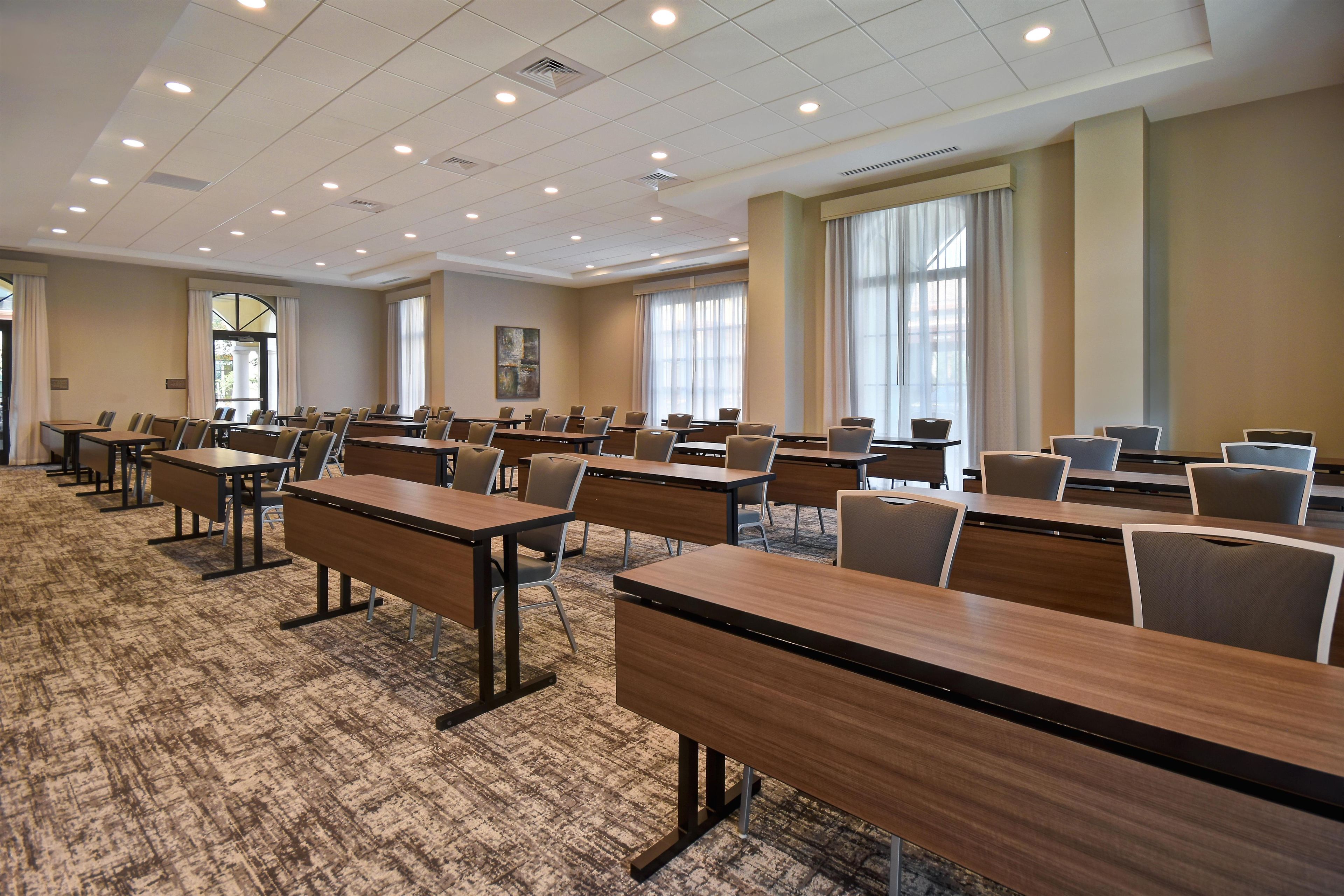A conference room with multiple tables and chairs, carpeted flooring, and large windows with curtains.