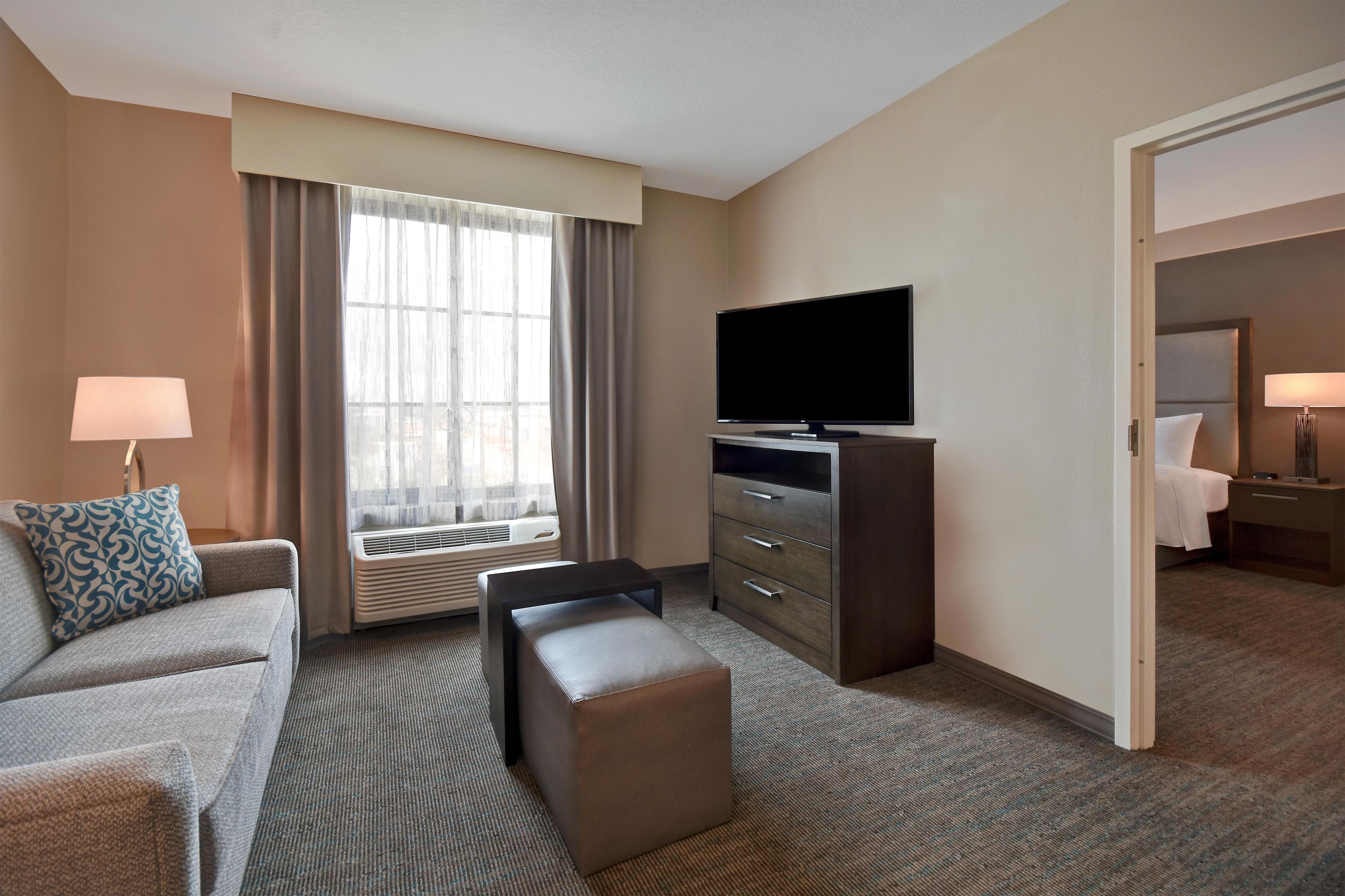 A hotel room with a flat-screen TV, a sofa, a small table, a dresser, and a window with curtains.