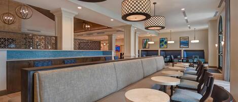 A modern restaurant with a long counter, cushioned seating, round tables, and patterned pendant lighting.