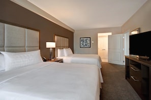 A hotel room with two beds, a desk with a lamp, a TV, and a door leading to another room.