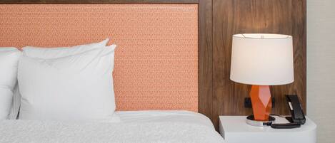 Una cama bien hecha con ropa blanca, un cabecero de madera con un panel naranja texturizado y una mesita de noche con una lámpara y un teléfono.