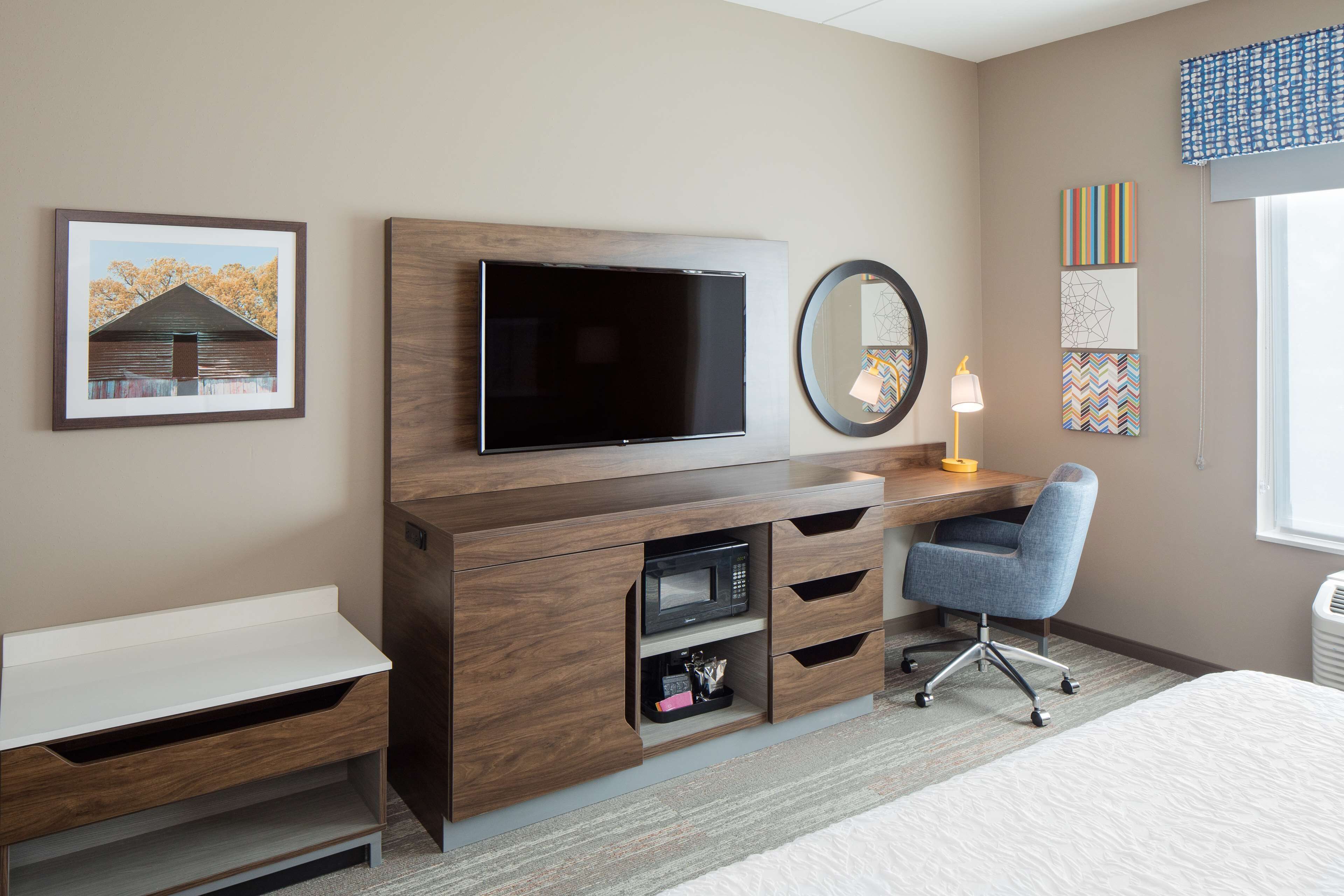 Habitación de hotel con televisión, escritorio con silla, cama, mesita de noche, espejo y obras de arte en las paredes.