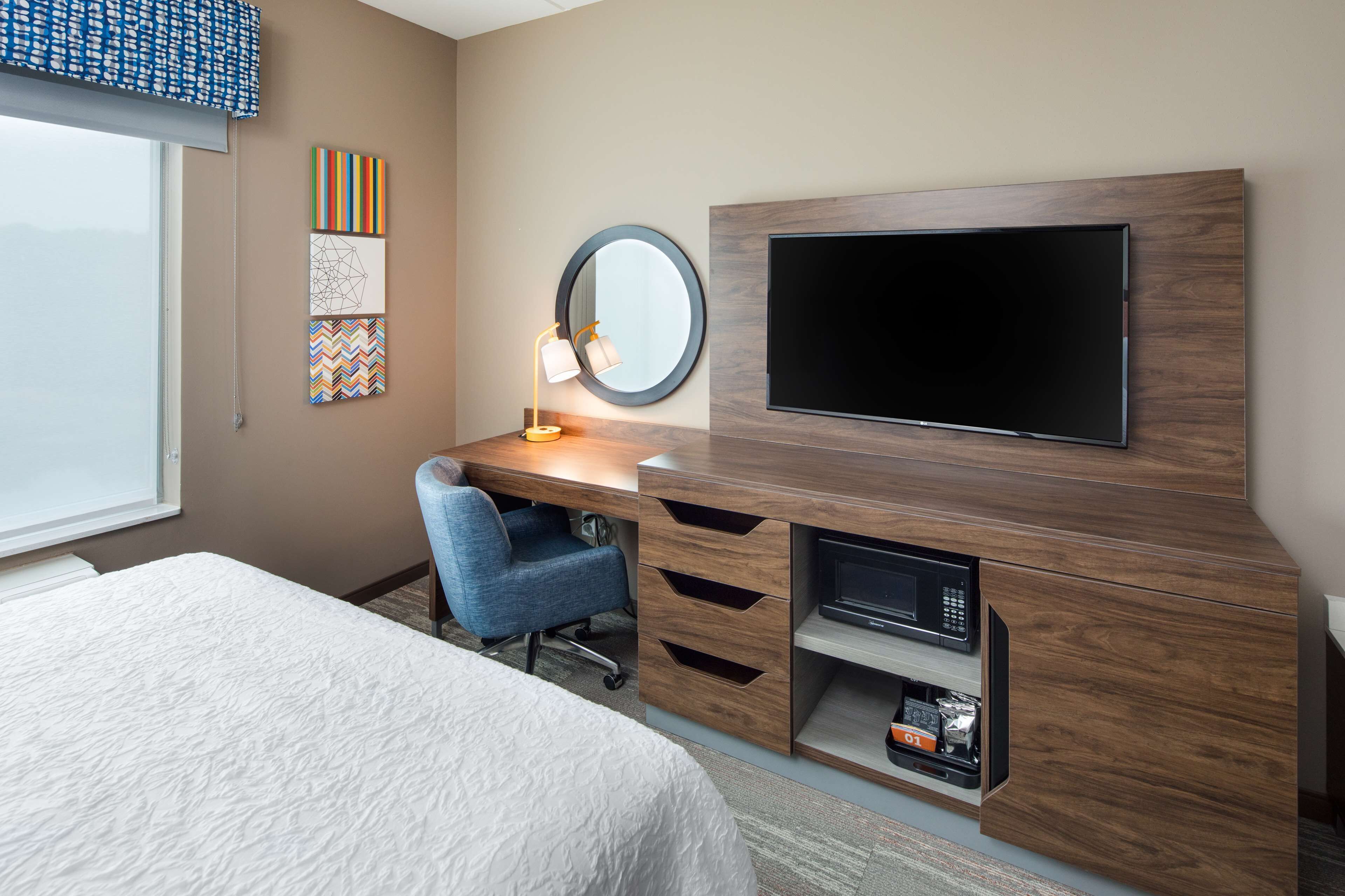 Habitación de hotel con cama, escritorio con silla, televisión, espejo y obras de arte en la pared.