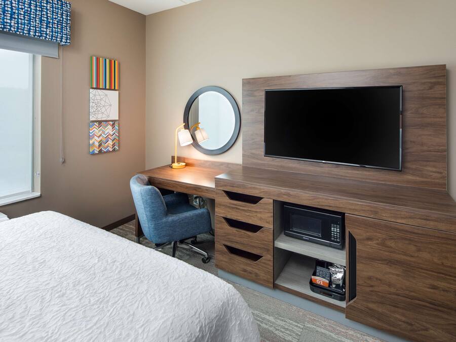 Habitación de hotel con cama, escritorio con silla, televisión, espejo y obras de arte en la pared.