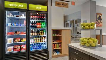 A hotel lobby with two beverage refrigerators labeled "really cold" and "cold," stocked with various drinks and snacks. There is a sign for "Home 2 Mkt" and an exit sign above a door. A bowl of green apples is on a counter.