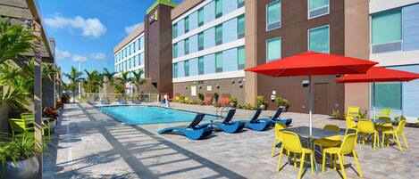 A hotel pool area with sun loungers and umbrellas, a swimming pool, and a building with multiple windows.