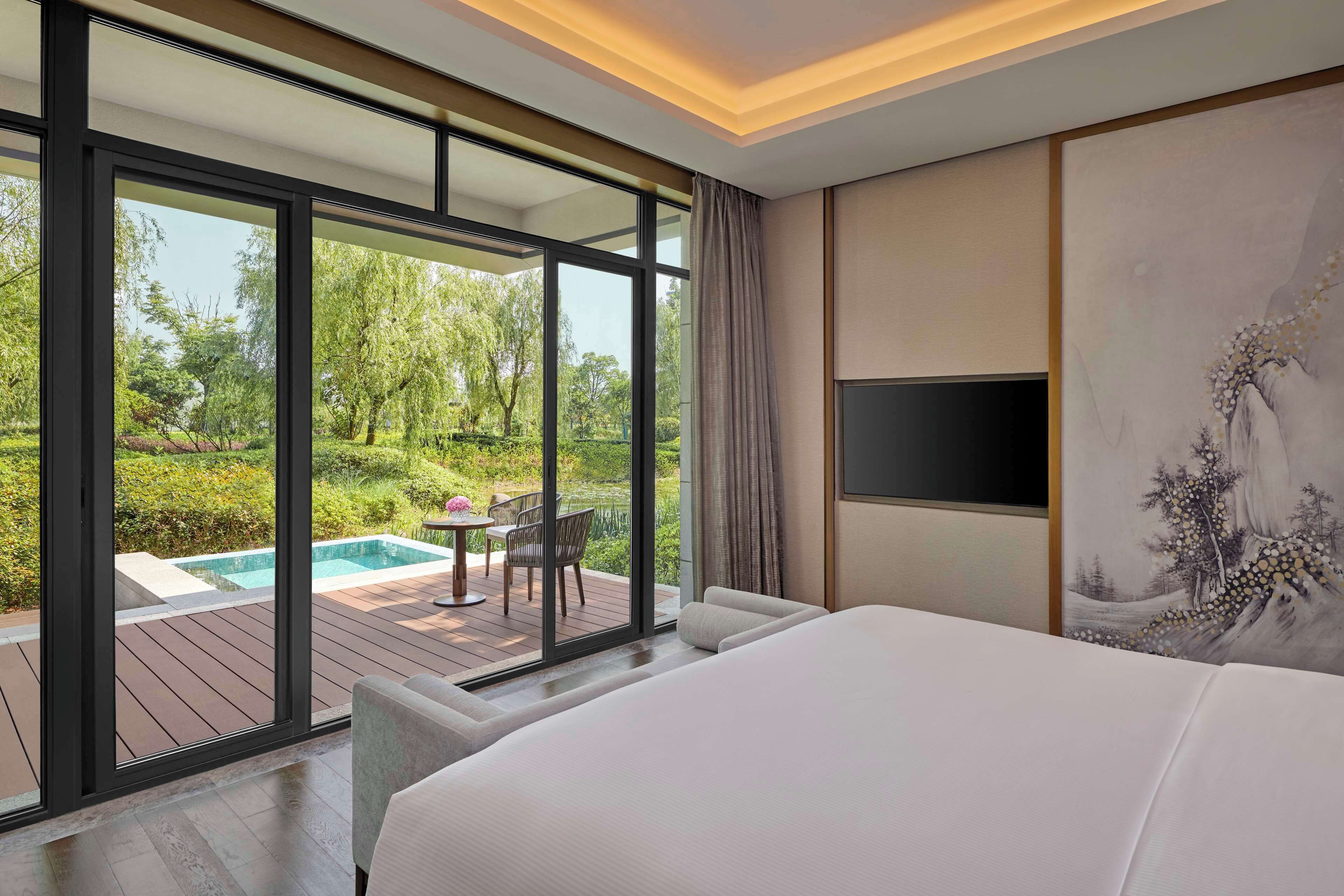 A modern hotel room with a large bed, a TV, and a view of an outdoor pool and garden.
