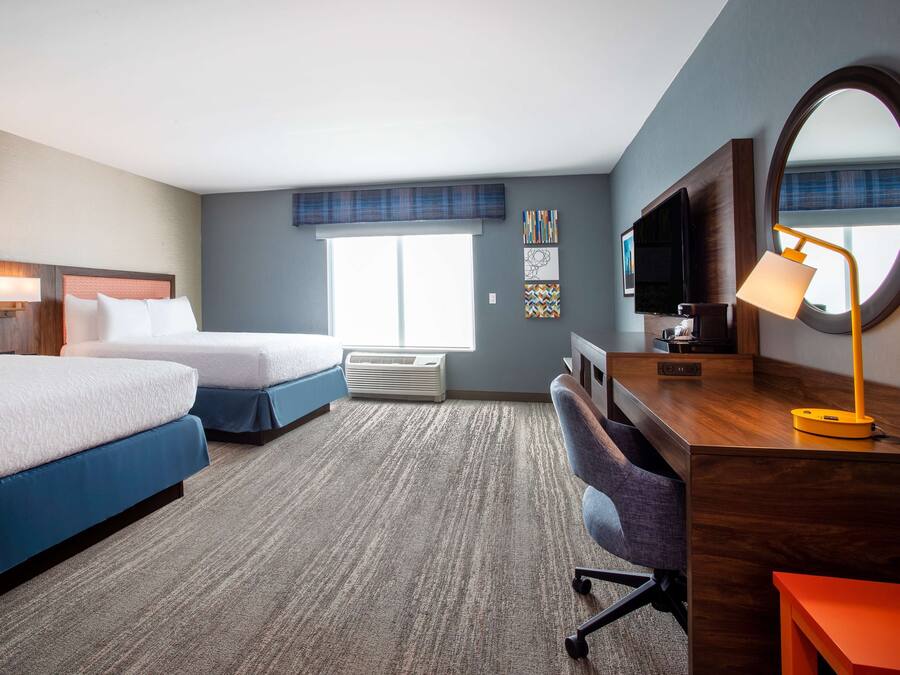 A hotel room with two beds, a desk with a chair, a TV, a lamp, and a large window.