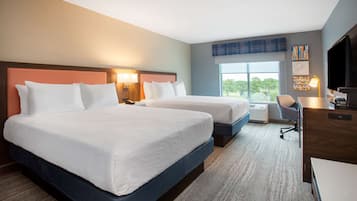 A hotel room with two beds, a desk, a chair, a television, and a large window with a view of trees.