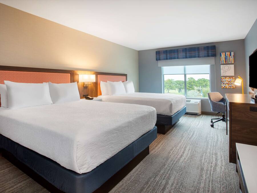 A hotel room with two beds, a desk, a chair, a television, and a large window with a view of trees.