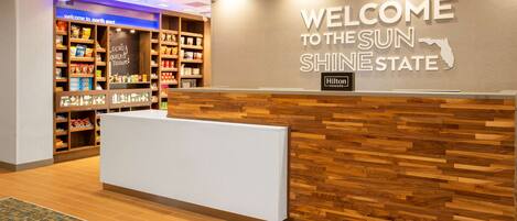 A hotel reception area with a wooden reception desk, a wall sign welcoming guests to the Sun Shire State, and a display of locally sourced treasures.