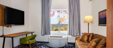 A hotel room with a flat-screen TV, a desk with a chair, a sofa, a small table, and a window with curtains.