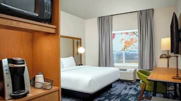 Habitación de hotel con cama, escritorio, silla, cafetera, televisor y ventana con cortinas.