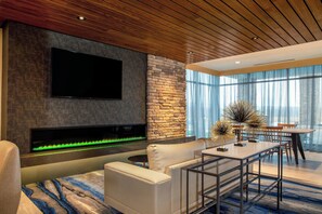 Una sala moderna con televisor de pantalla plana, una pared de acento de piedra, chimenea con fuego iluminado en verde, un sofá, una mesa de centro y un comedor con mesa y sillas.