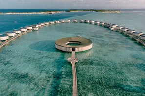 An aerial view of a resort with a circular building surrounded by overwater bungalows connected by a pier.