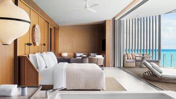 Una habitación de hotel moderna con una cama grande, un área de descanso con vista al océano, un ventilador de techo y un ventanal amplio.