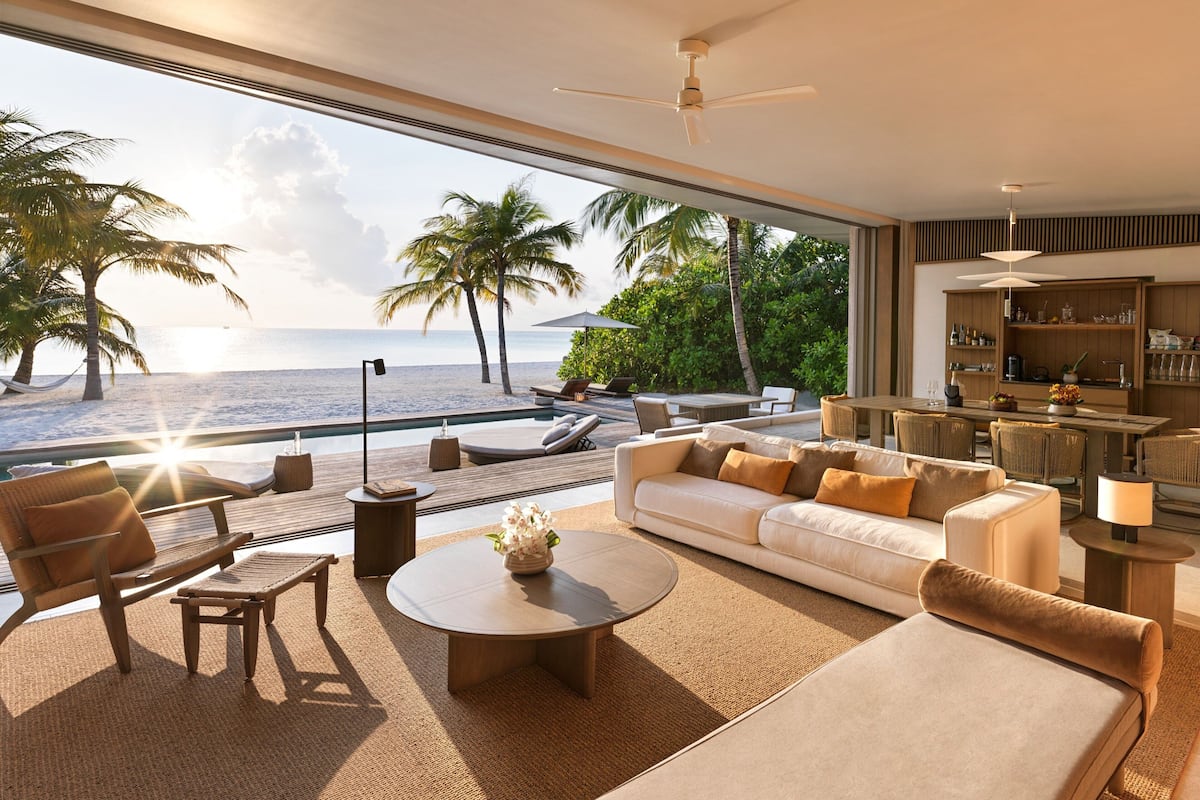 A modern living room with a large window overlooking a beach, furnished with a sofa, chairs, and a coffee table.