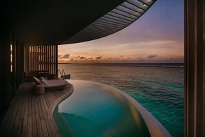 A modern infinity pool with a wooden deck and lounge chairs, overlooking a calm sea at sunset.