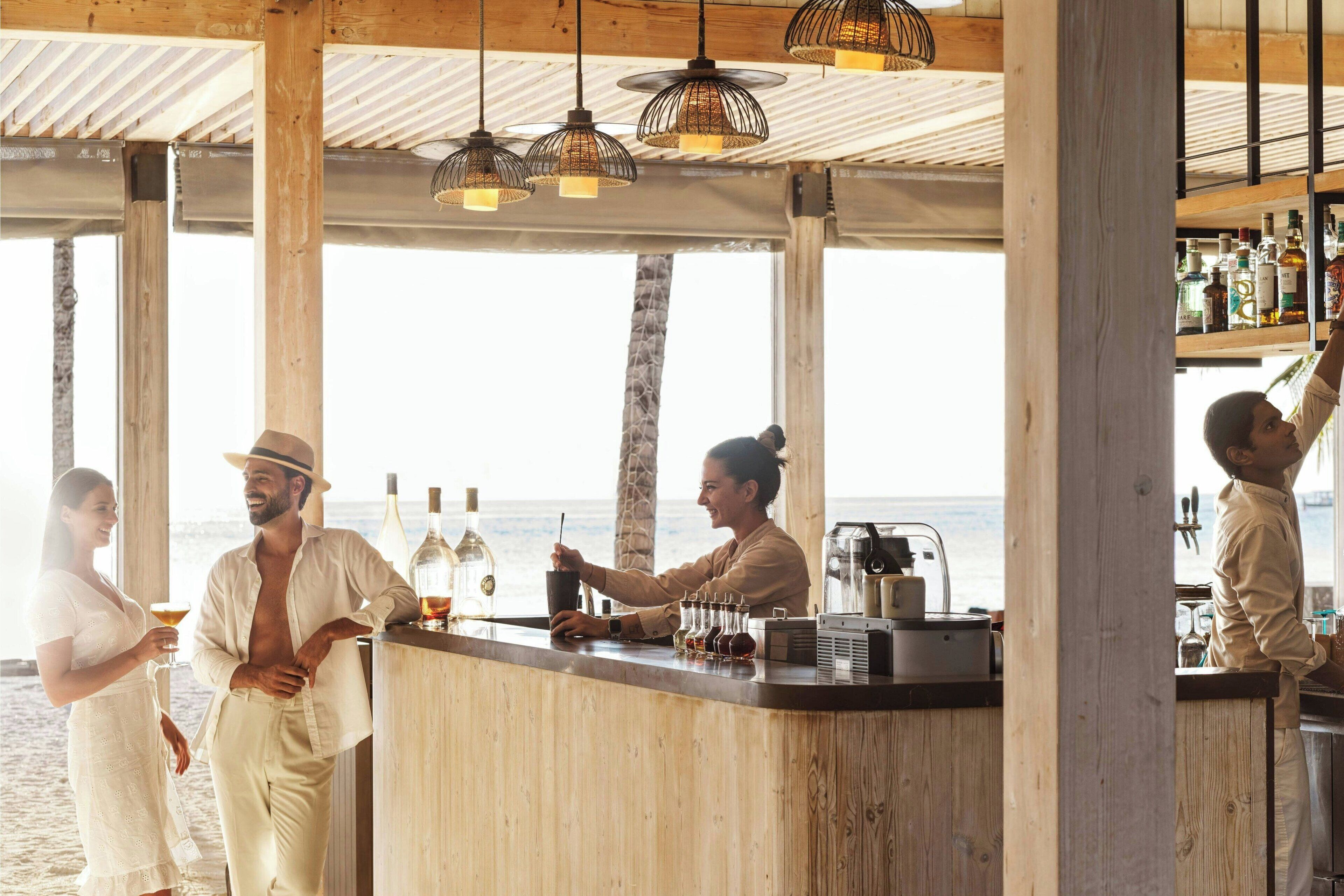 Een strandbar met houten balken, hangende lampen en uitzicht op de oceaan.