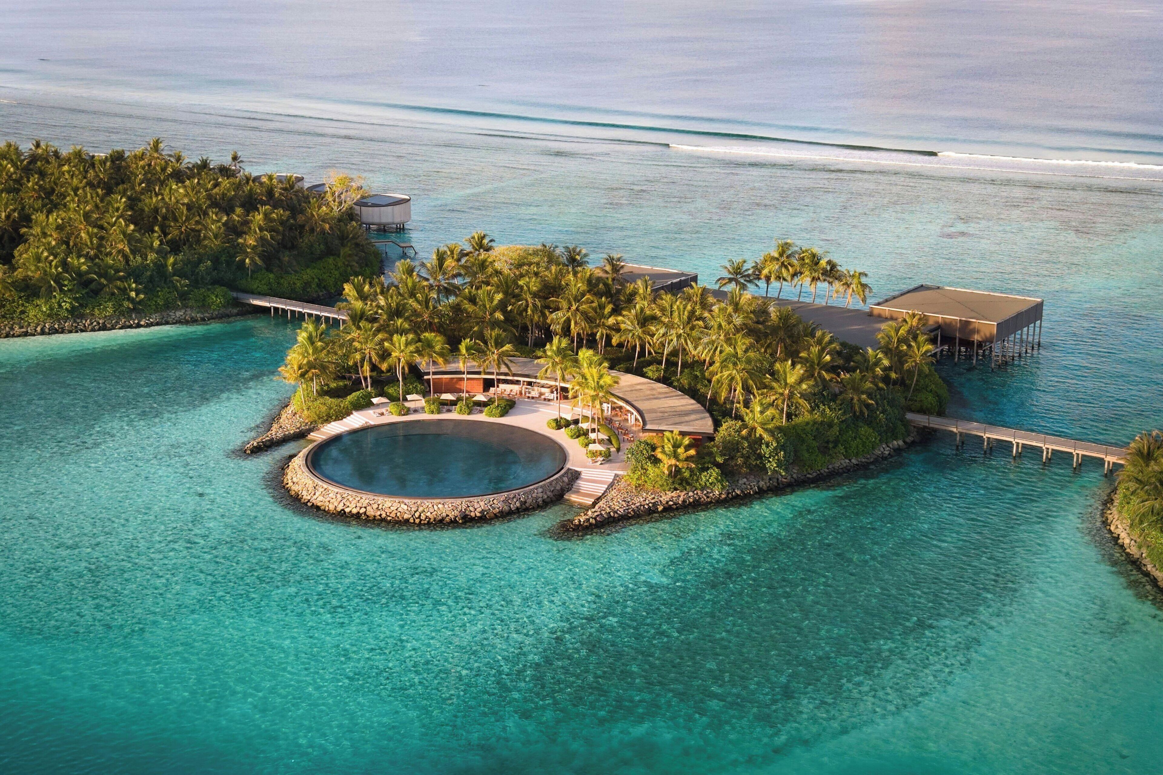 Vue aérienne d’une île-station tropicale avec une piscine circulaire, des palmiers et une jetée.