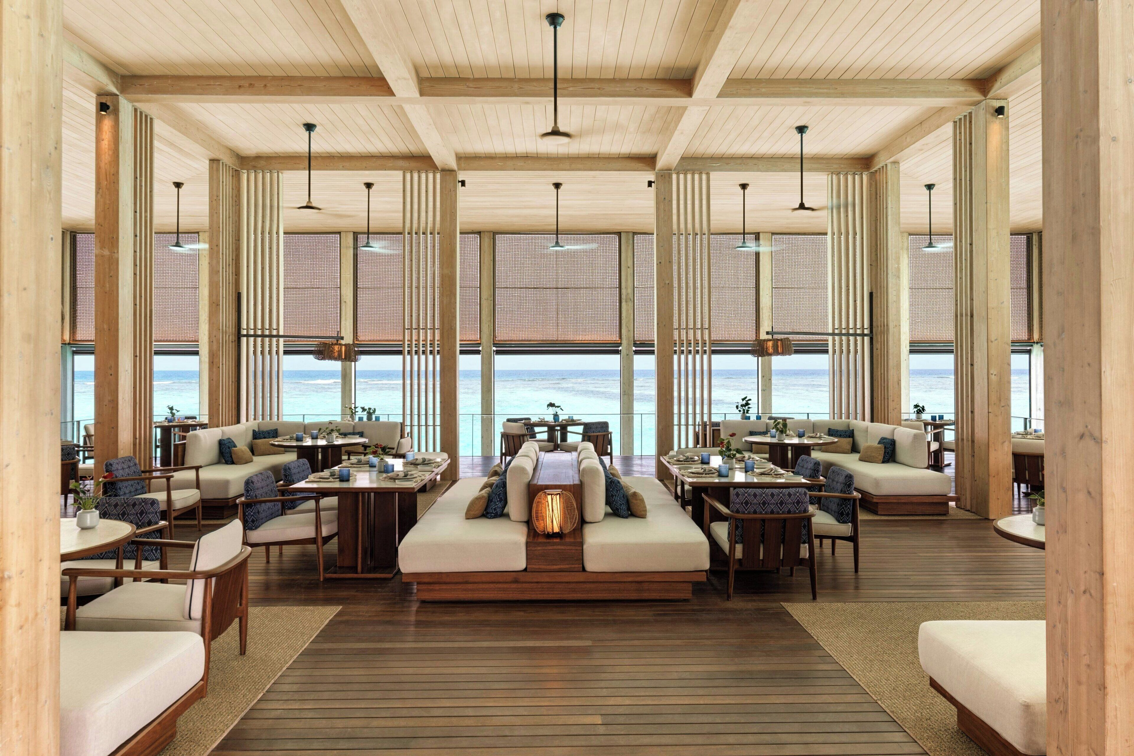 Un espacio interior amplio con piso de madera, varios conjuntos de asientos y ventanas grandes con vistas al mar.
