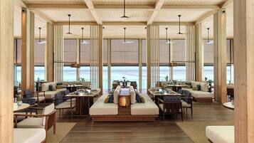Un espacio interior amplio con piso de madera, varios conjuntos de asientos y ventanas grandes con vistas al mar.