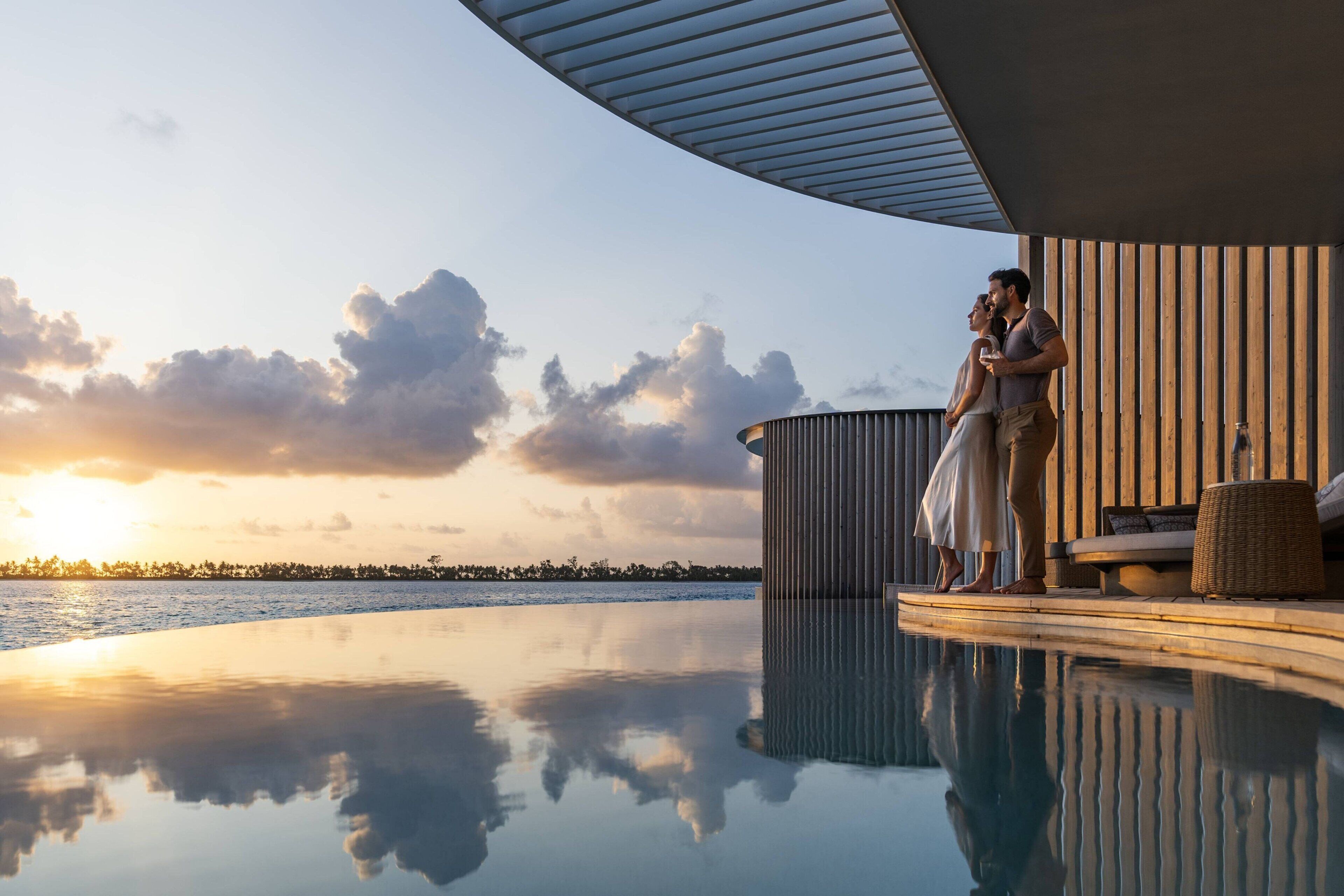 A couple standing by a pool with a sunset view, wooden deck, and lounge area.