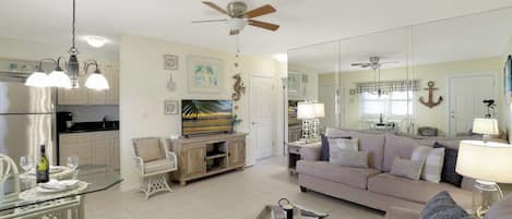 A spacious living room with a sofa, a glass coffee table, and a television. There is a kitchen area with cabinets and a refrigerator.