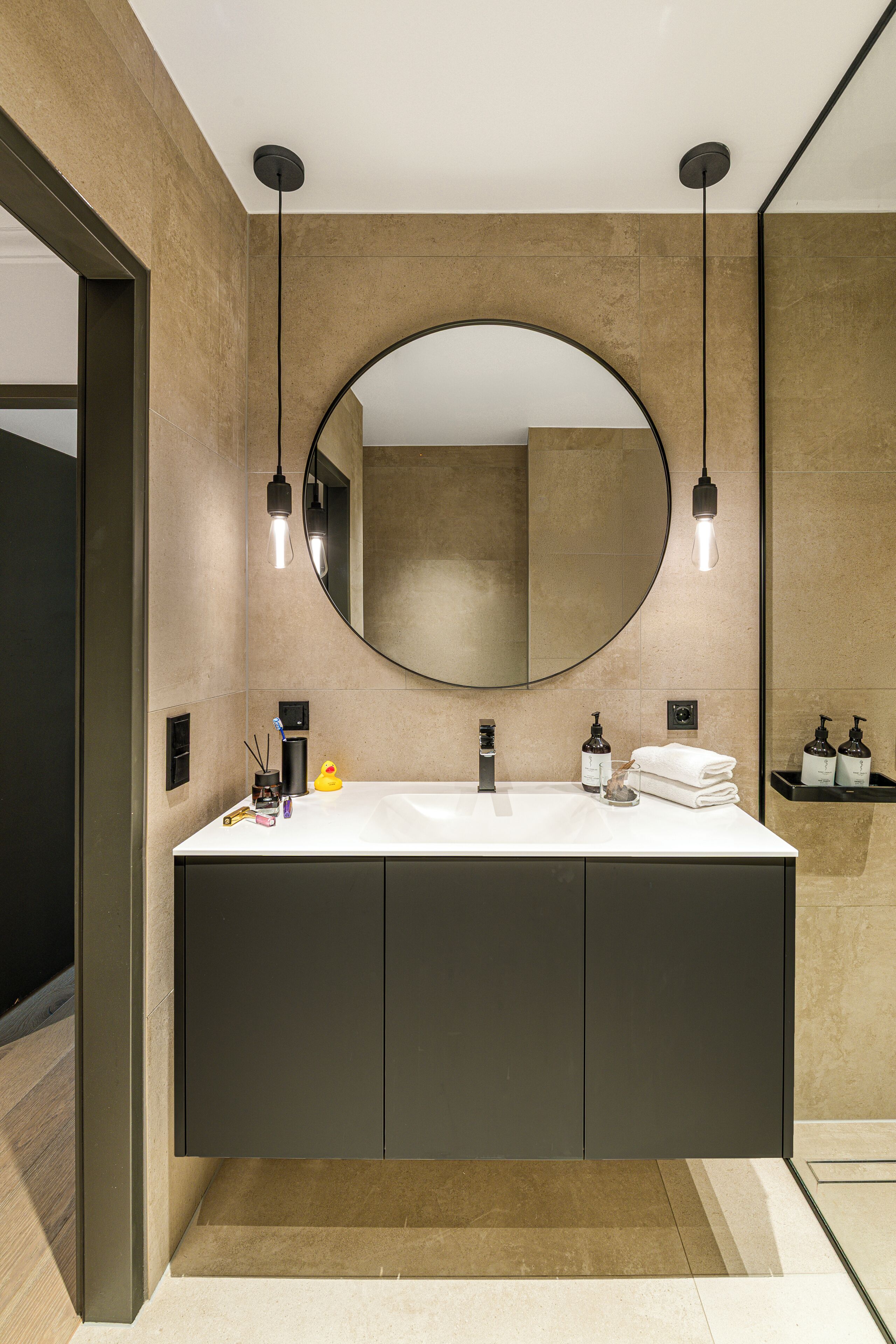 Un baño moderno con un espejo grande, dos lámparas colgantes, un lavabo blanco y un mueble oscuro.