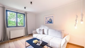 A modern living room with a sofa, a coffee table with books and a teapot, a window with curtains, and a framed picture on the wall.