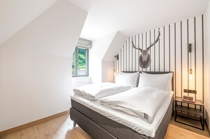 Um quarto com uma cama, uma decoração de cabeça de veado fixada na parede, uma mesa de cabeceira com livros e uma janela com vista para o verde.