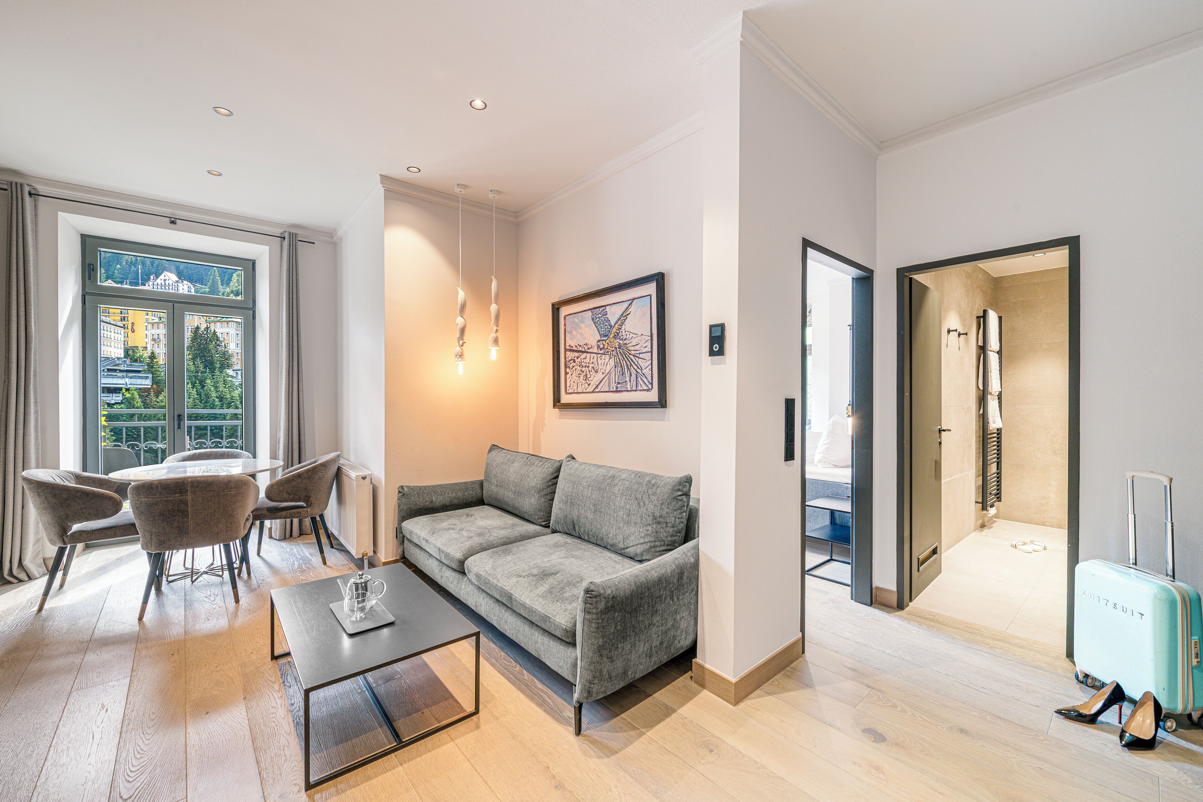 Une chambre d’hôtel moderne avec un canapé gris, une table à manger et des chaises, une grande fenêtre avec des rideaux et une vue sur les bâtiments.