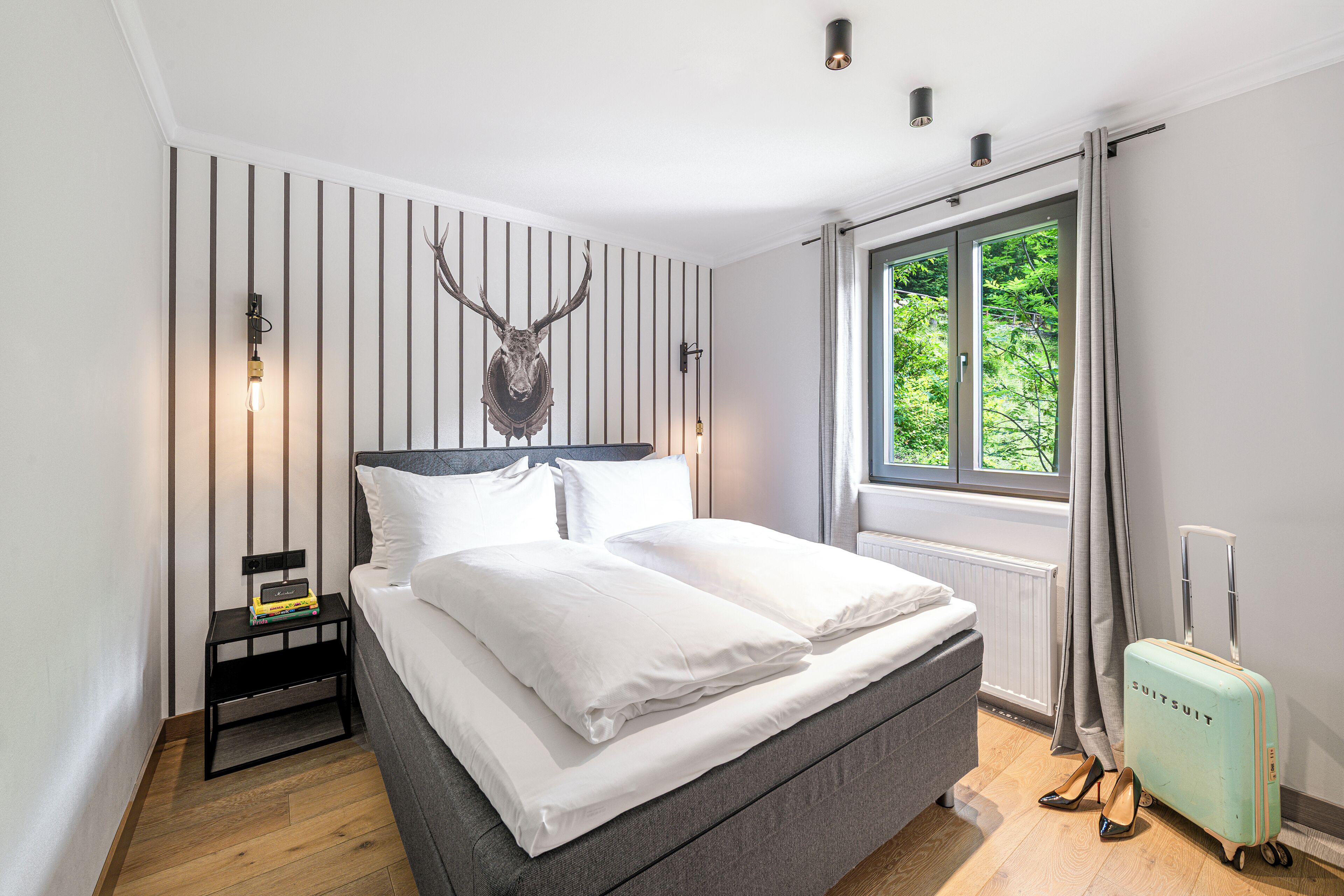 A bedroom with a bed, a mounted deer head, a window with curtains, a radiator, and a suitcase.