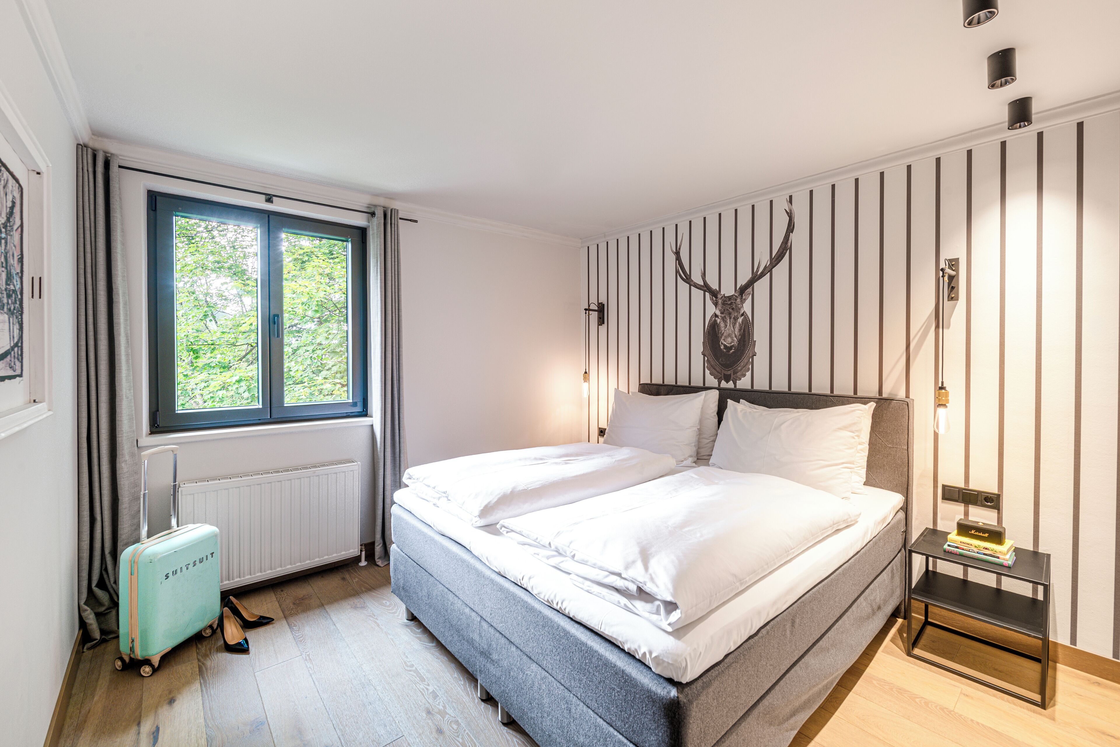 A hotel room with a bed, a suitcase, a radiator, a window with curtains, and a wall-mounted deer head decoration.