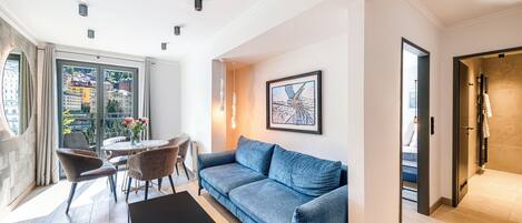 Una sala de estar moderna con un sofá azul, una mesa de centro y un comedor con sillas. Hay un cuadro en la pared y vistas de edificios a través de un ventanal grande.