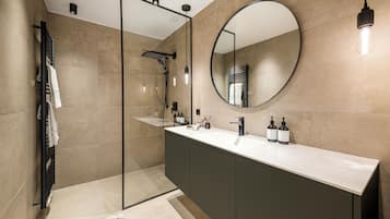 A modern bathroom with a glass-enclosed shower, a round mirror, and a vanity with a sink and two bottles of shampoo.