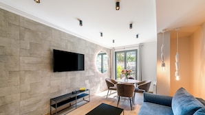 A modern living room with a flat-screen TV mounted on the wall, a sofa, a coffee table, and a dining area with chairs and a table.