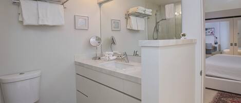 A modern bathroom with a toilet, sink, and mirror. There are towels hanging on a rack and a shower visible in the mirror.