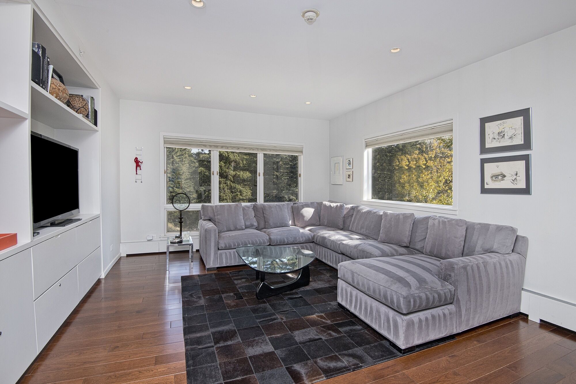 A modern living room with a sectional sofa, a glass coffee table, and a flat-screen TV. There are large windows providing a view of trees outside.