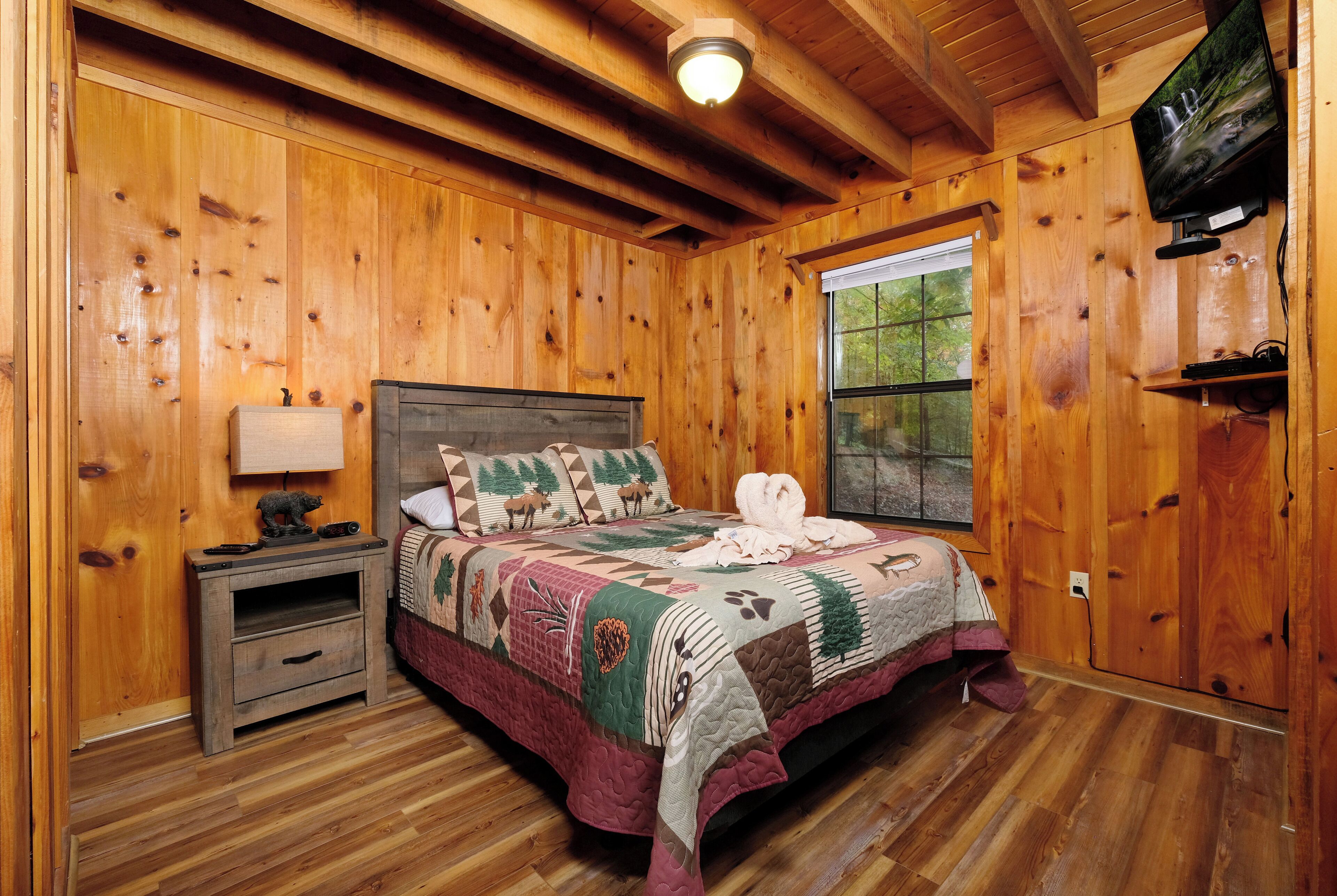 A wooden cabin bedroom with a bed, a nightstand, a television, and a window.