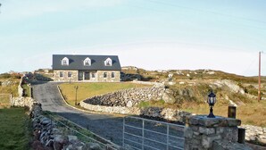 A stone house with a dark roof, a stone wall in front, and a paved road leading to it. - Cnocan Na Cuaigh Carraroe (Carraroe)