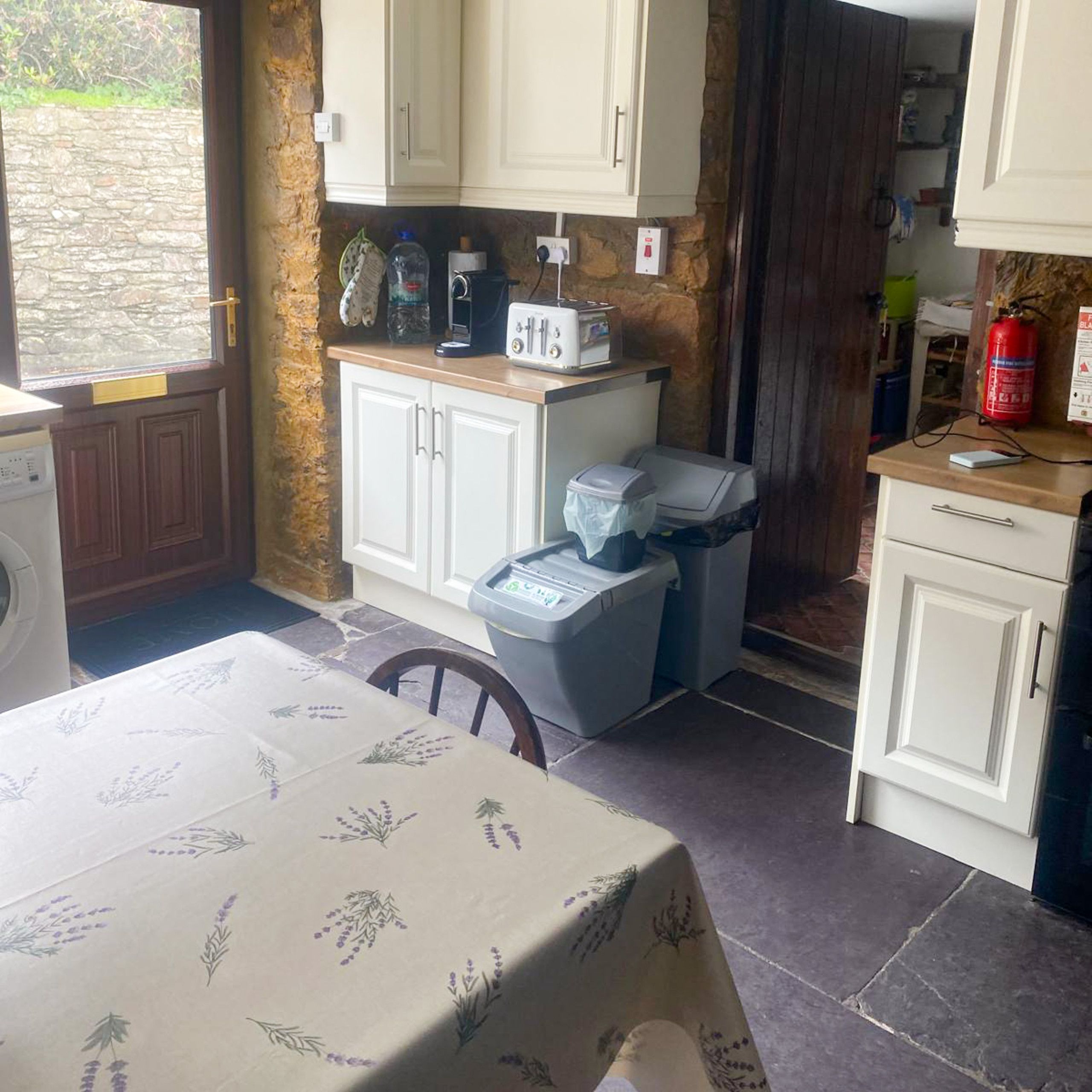 A kitchen with white cabinets, a table with a floral tablecloth, a washing machine, and a fire extinguisher.